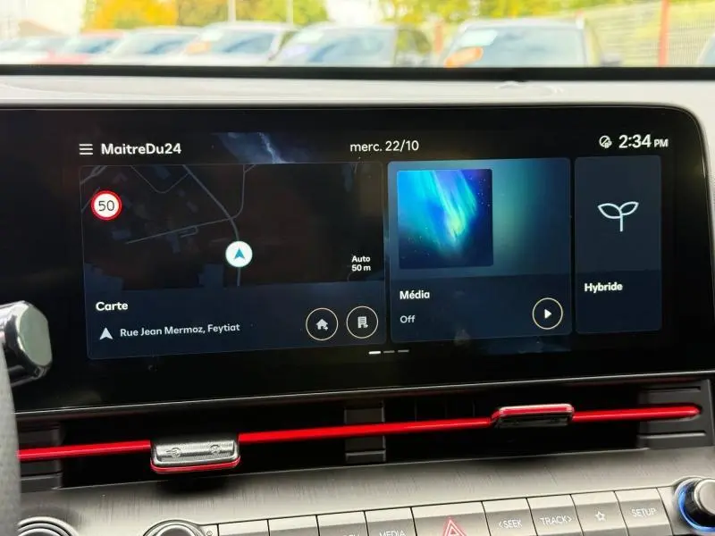 Écran tactile central du Hyundai Kona 2025 affichant navigation, média et mode hybride, avec aérateurs rouges en dessous.