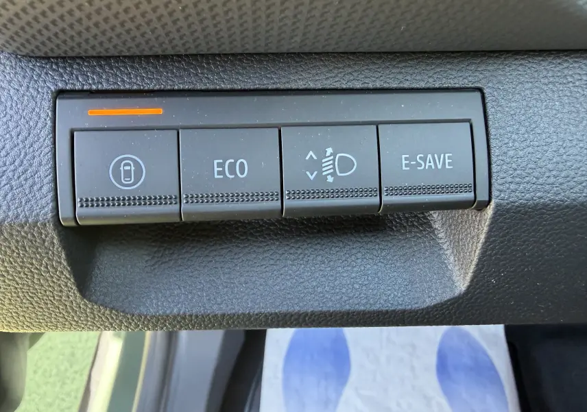 Gros plan sur les boutons de commande du tableau de bord du Dacia Duster III kaki, avec témoin orange allumé.