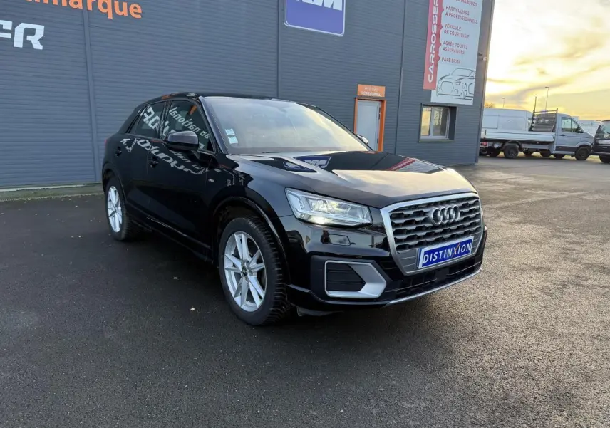 Audi Q2 S Line noir brillant vue 3/4 avant droit sur parking devant bâtiment commercial.