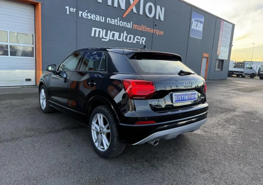 Audi Q2 noir brillant vue 3/4 arrière droit, avec feux rouges et jantes argentées visibles devant un bâtiment commercial.