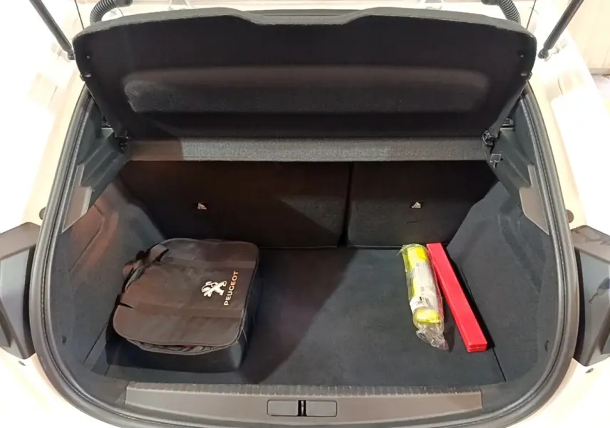 Coffre ouvert d'une Peugeot 208 électrique blanche, montrant un sac Peugeot noir et deux objets de sécurité à l'intérieur.
