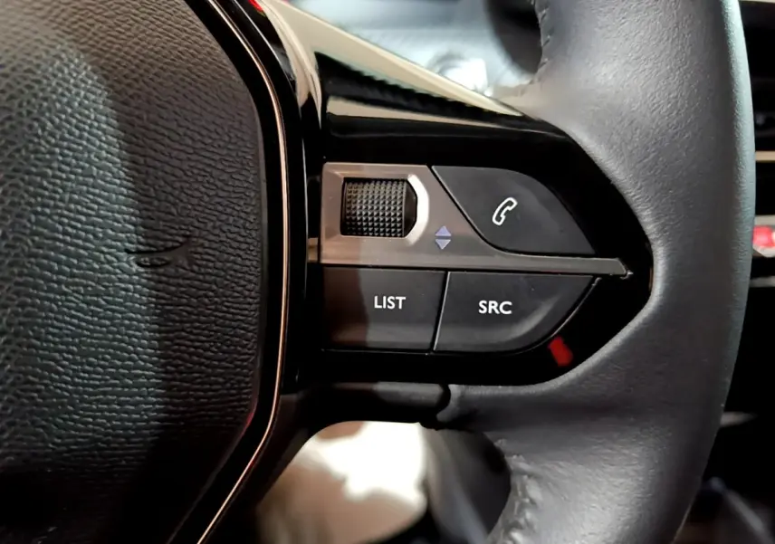 Gros plan sur les commandes gauche du volant cuir noir de la Peugeot 208 électrique blanche, boutons LIST et SRC visibles.