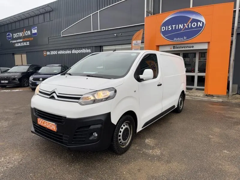 Citroën Jumpy blanc banquise en 3/4 avant droit, avec calandre noire et feux allumés, stationné devant un garage.