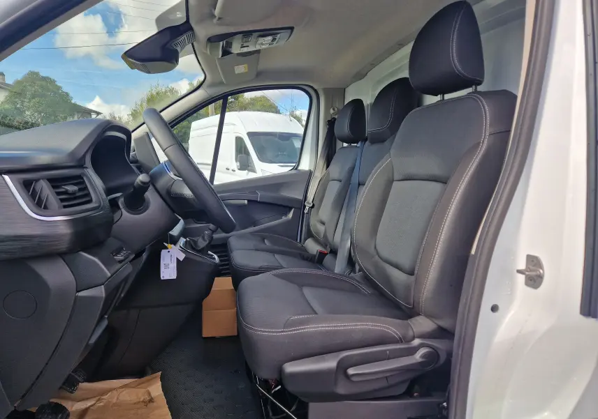 Vue intérieure côté conducteur du Nissan Primastar blanc 2025, montrant les trois sièges avant en tissu noir et le tableau de bord.