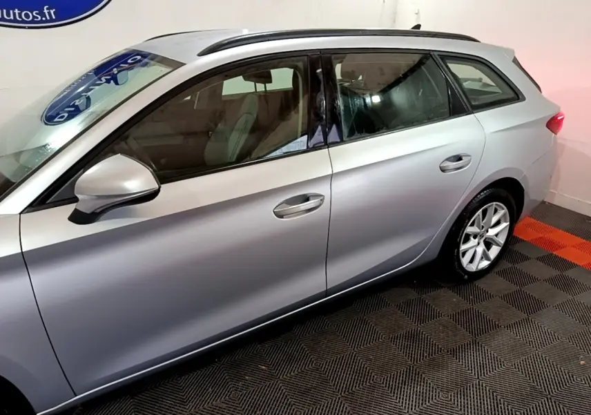 Vue latérale côté gauche d'une SEAT LEON gris clair 2022 en stationnement intérieur sur sol damier noir et orange.