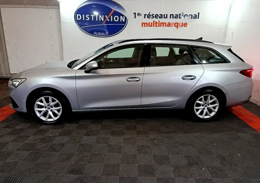 Profil côté gauche d'une SEAT LEON gris clair break 2022, roues alliage et rails de toit noirs visibles en intérieur showroom.