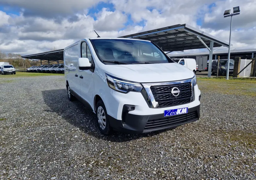 Vue 3/4 avant droit d’un utilitaire Nissan Primastar blanc avec calandre noire et logo Nissan visible, sur parking extérieur.
