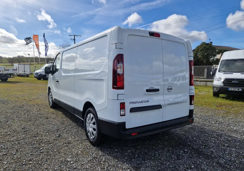 Vue 3/4 arrière droite d'un utilitaire Nissan Primastar blanc, avec portes arrière fermées sur un terrain extérieur.