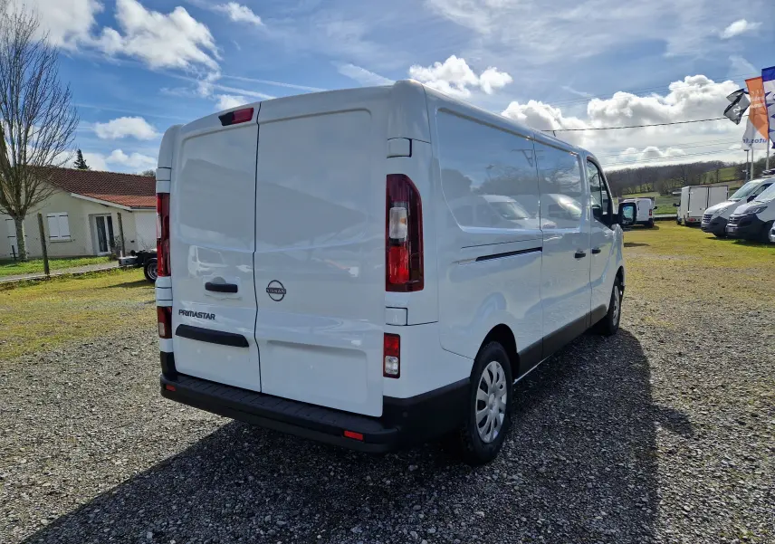 Vue 3/4 arrière droite d’un utilitaire Nissan Primastar blanc avec portes arrière battantes sur un terrain extérieur.