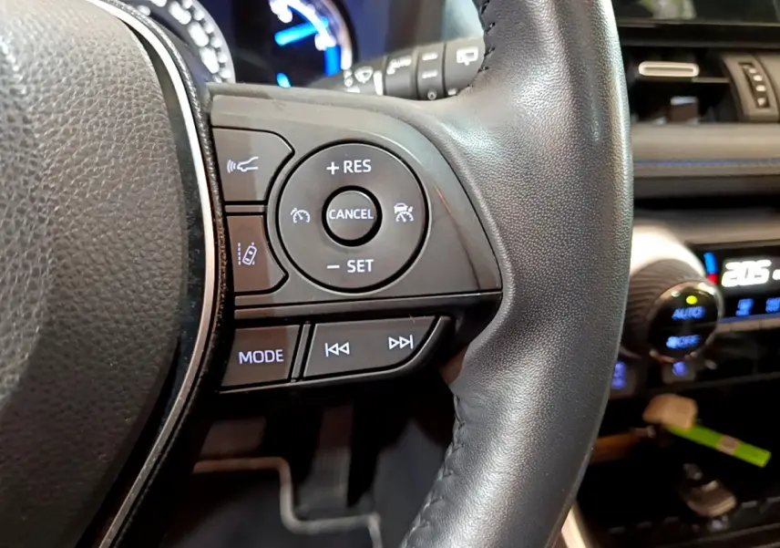 Gros plan sur les commandes du volant en cuir noir du Toyota RAV4 2WD 218ch Collection, avec tableau de bord lumineux en arrière-plan.