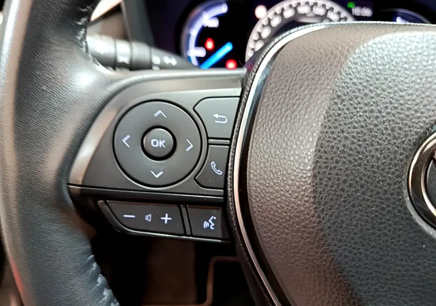 Gros plan sur les commandes à gauche du volant cuir noir du Toyota RAV4 gris clair, tableau de bord lumineux en arrière-plan.