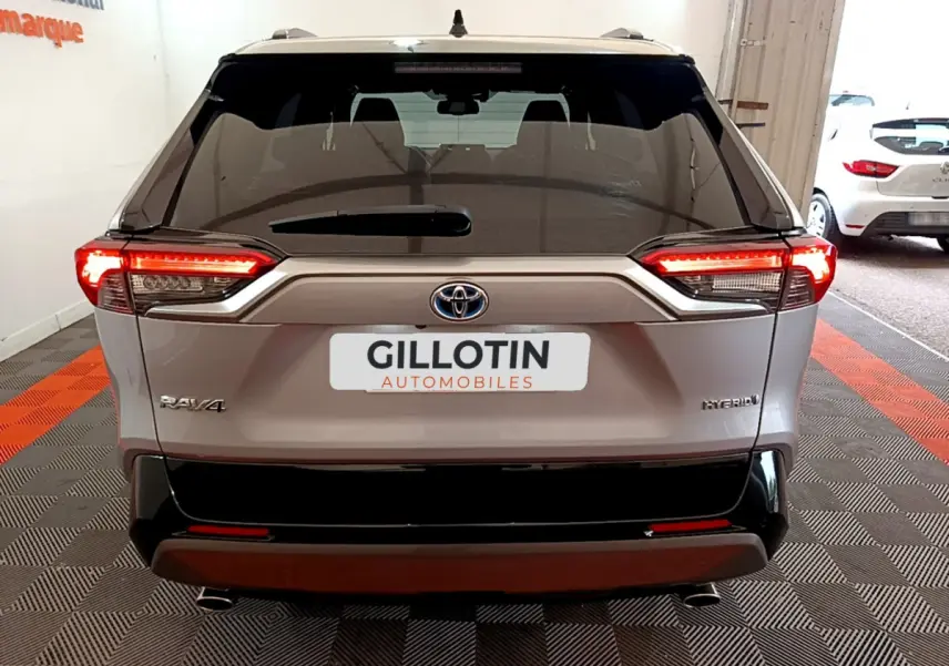 Vue arrière du Toyota RAV4 gris clair hybride avec feux LED allumés et logo visible sur un sol à damiers rouge et noir.