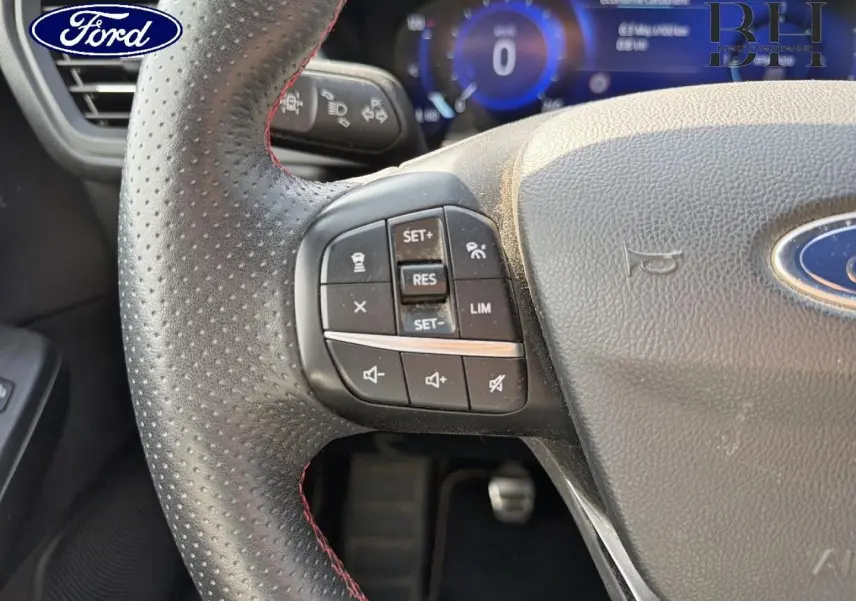 Gros plan sur le volant cuir perforé du Ford Kuga bleu, avec commandes multifonctions et tableau de bord numérique en arrière-plan.