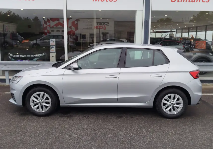 Vue de profil côté gauche d'une Skoda Fabia gris argent métallisé 2023 garée devant une vitrine de concession.