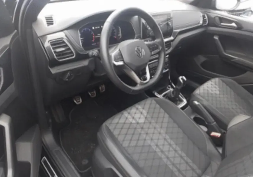 Vue intérieure côté conducteur du Volkswagen T-Cross 2025 noir métal, avec volant multifonction, sièges tissu à motif losanges et levier de vitesse manuel.