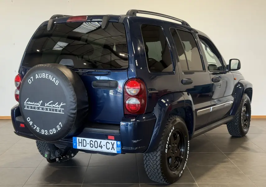 Jeep Cherokee bleu vue 3/4 arrière droit, avec roue de secours couverte et jantes noires tout-terrain.