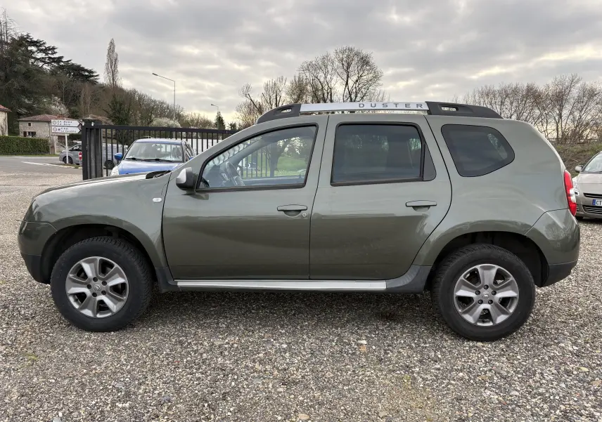 Vue de profil côté gauche d'un Dacia Duster 1.5 DCI 110 vert foncé avec barres de toit et jantes noires sur un sol gravillonné.
