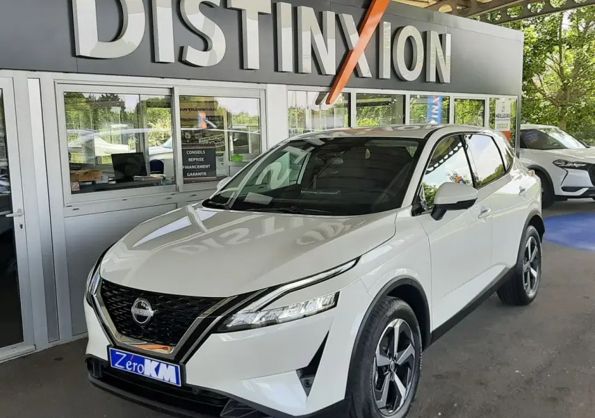 Nissan Qashqai blanc lunaire en 3/4 avant droit, avec calandre noire et jantes alliage distinctives, sous un auvent showroom.