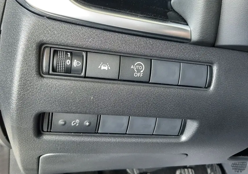 Vue rapprochée des commandes de réglage d'éclairage et d'assistance sur la gauche du tableau de bord d'un Nissan Qashqai blanc lunaire 2022.