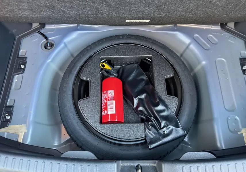 Vue plongeante sur la roue de secours noire et un extincteur rouge dans le coffre d'une Toyota Yaris Hybride bleu métal.