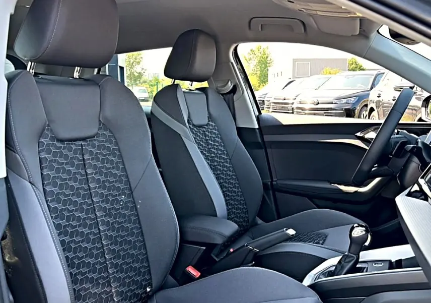 Vue intérieure côté avant droit de l’Audi A1 Sportback 2026, sièges sport gris avec motifs hexagonaux et accoudoir central.