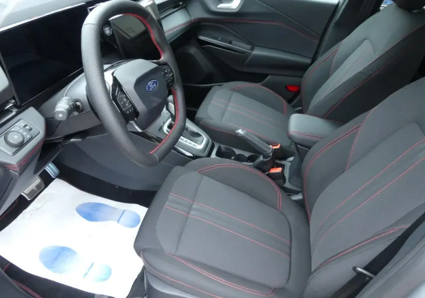 Intérieur avant du Ford Puma 2025, sièges en tissu gris avec surpiqûres rouges et volant sport multifonction.
