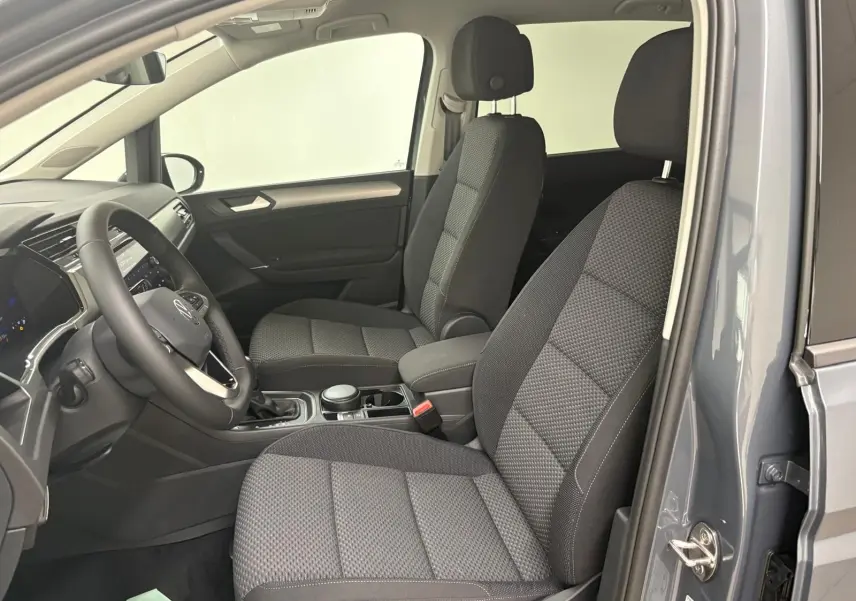 Vue intérieure côté conducteur du Volkswagen Touran gris dauphin 2025, montrant les sièges avant en tissu et le tableau de bord.