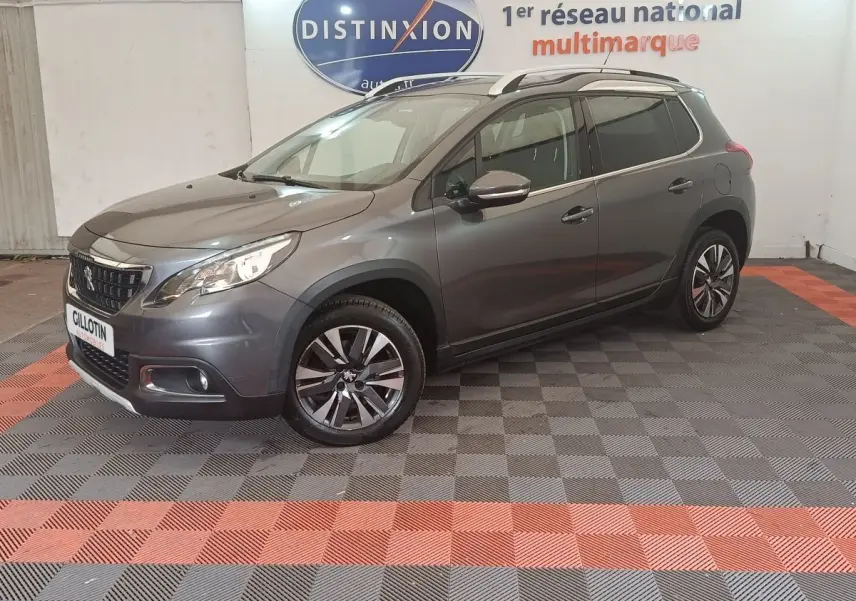 Peugeot 2008 gris foncé en 3/4 avant droit, avec jantes alliage et barres de toit chromées visibles.