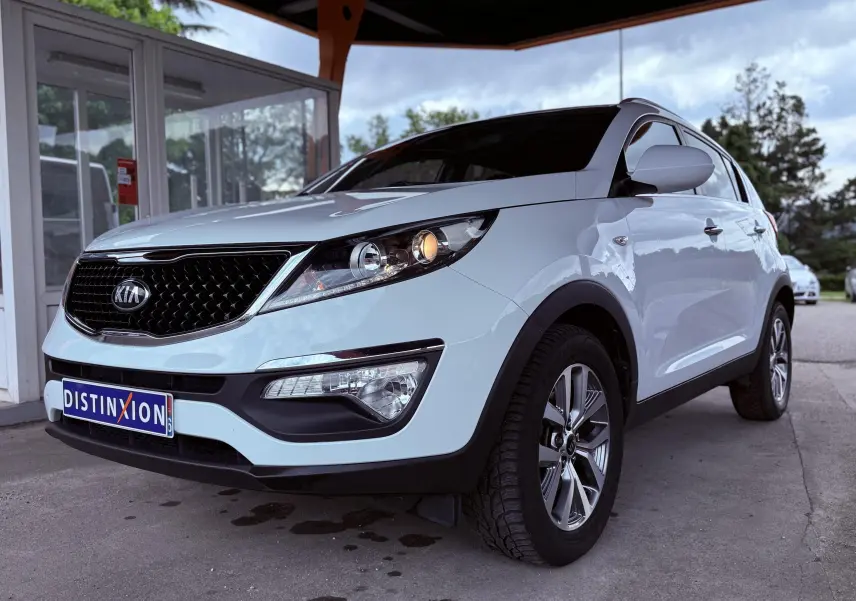 KIA Sportage blanche vue en 3/4 avant droit, avec calandre noire et phares allumés sous un abri.