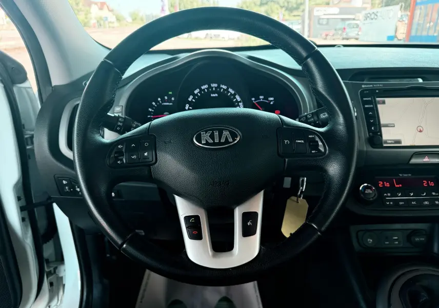 Vue intérieure centrée sur le volant noir du Kia Sportage blanc de 2015 avec tableau de bord et écran GPS visible.