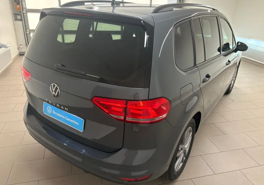 Vue 3/4 arrière droite d'un Volkswagen Touran gris dauphin 2025 avec feux arrière LED et hayon fermé en intérieur showroom.