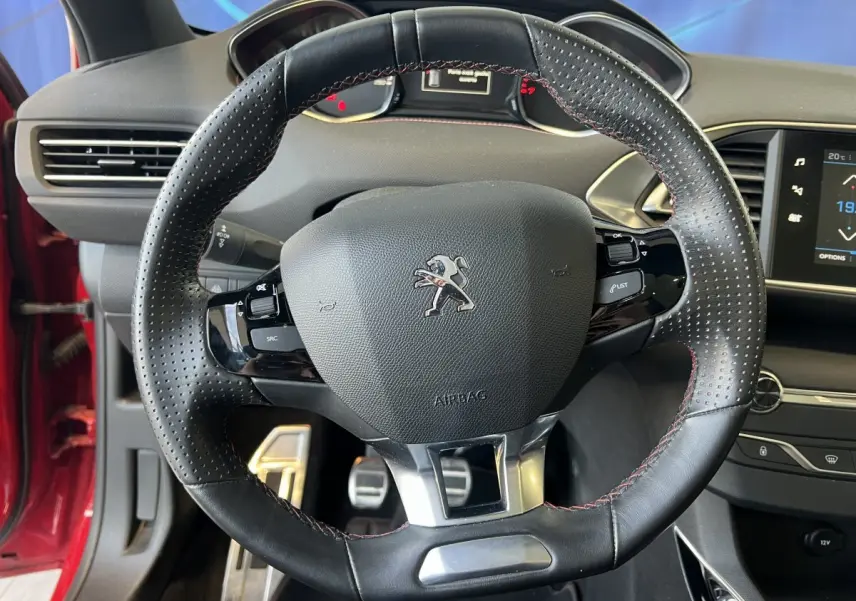 Vue rapprochée du volant cuir perforé avec coutures rouges de la Peugeot 308 GT Line rouge, tableau de bord en arrière-plan.