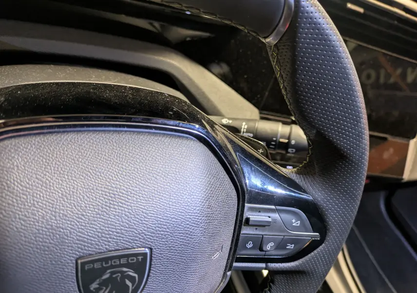 Gros plan sur le volant perforé noir de la Peugeot 308 SW Hybrid 145 eDCS6 GT, avec commandes intégrées visibles.