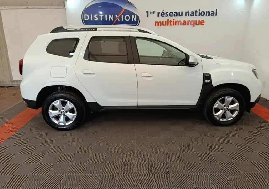 Vue de profil côté droit du Dacia Duster blanc Confort Blue dCi 115 4x4 avec rails de toit noirs en intérieur showroom.