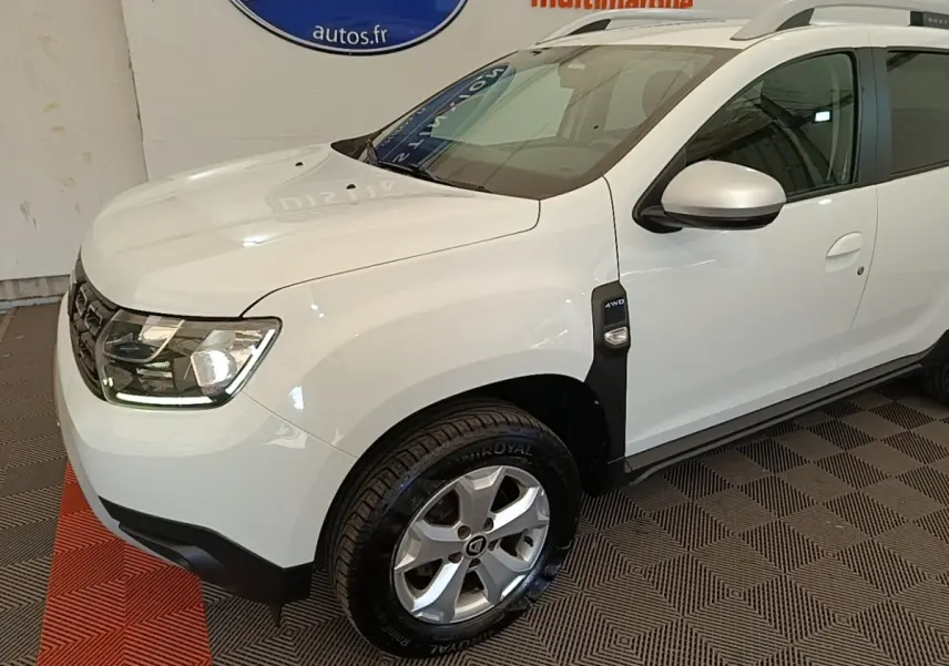 Vue 3/4 avant droit du Dacia Duster blanc avec badge 4x4 sur l'aile et jantes alliage visibles.