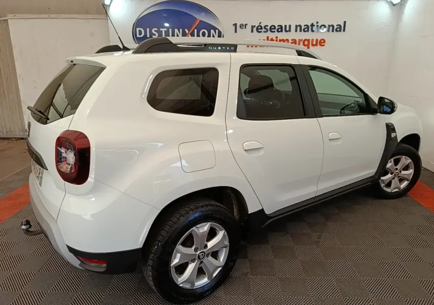 Vue 3/4 arrière droite d'un Dacia Duster blanc 2021 avec barres de toit noires et jantes alliage.