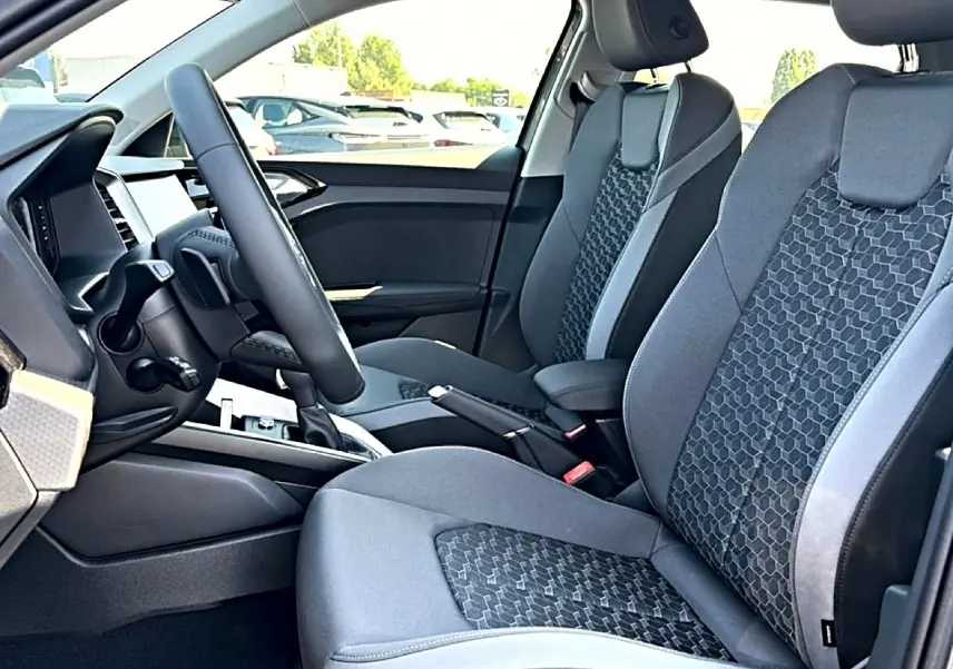 Vue intérieure côté conducteur de l'Audi A1 Sportback 2026, sièges sport gris avec motifs hexagonaux et volant cuir multifonction.