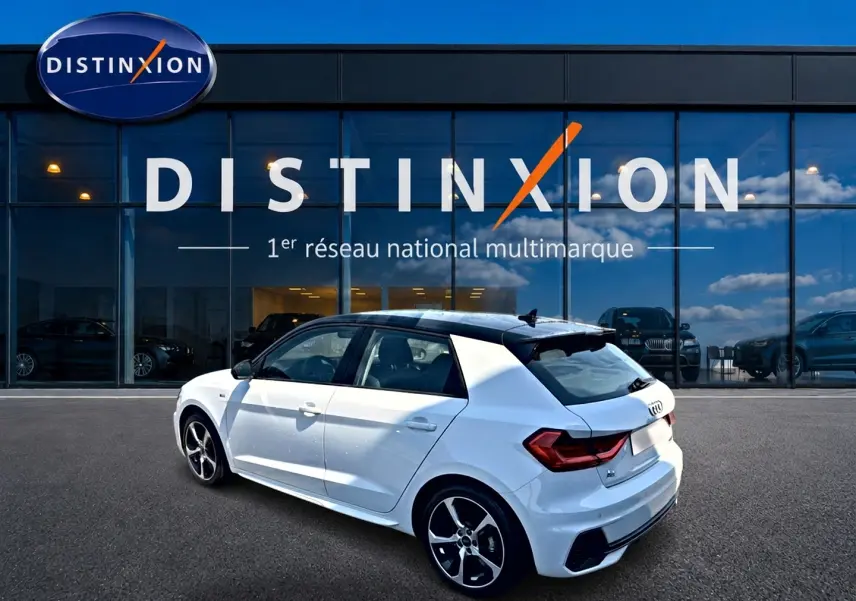 Audi A1 Sportback blanc avec toit noir en 3/4 arrière droit, jantes alu et becquet arrière visible devant showroom Distinxion.