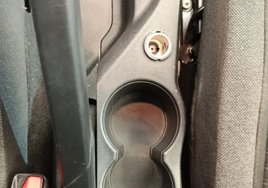 Gros plan sur la console centrale noire avec porte-gobelets et prise 12V entre les sièges avant gris foncé.