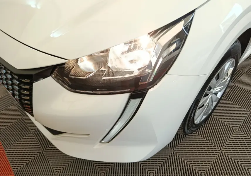 Gros plan sur le phare avant droit allumé d'une Peugeot 208 blanche, avec la calandre et la roue visibles.