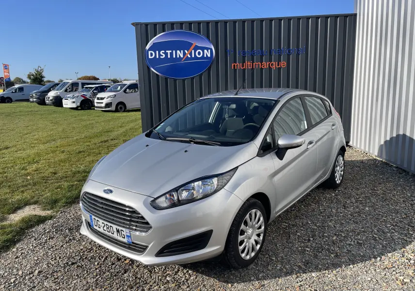 Ford Fiesta 1.25 82 CV Trend gris titane vue 3/4 avant droit sur parking gravillonné devant un mur noir.