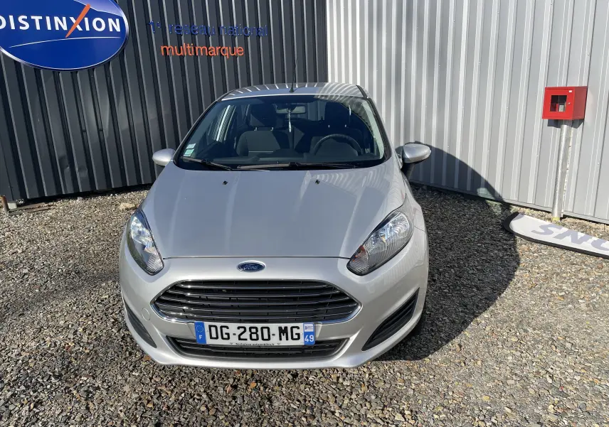 Vue frontale d'une Ford Fiesta gris titane 2014 stationnée devant un mur métallique avec plaque d'immatriculation française visible.