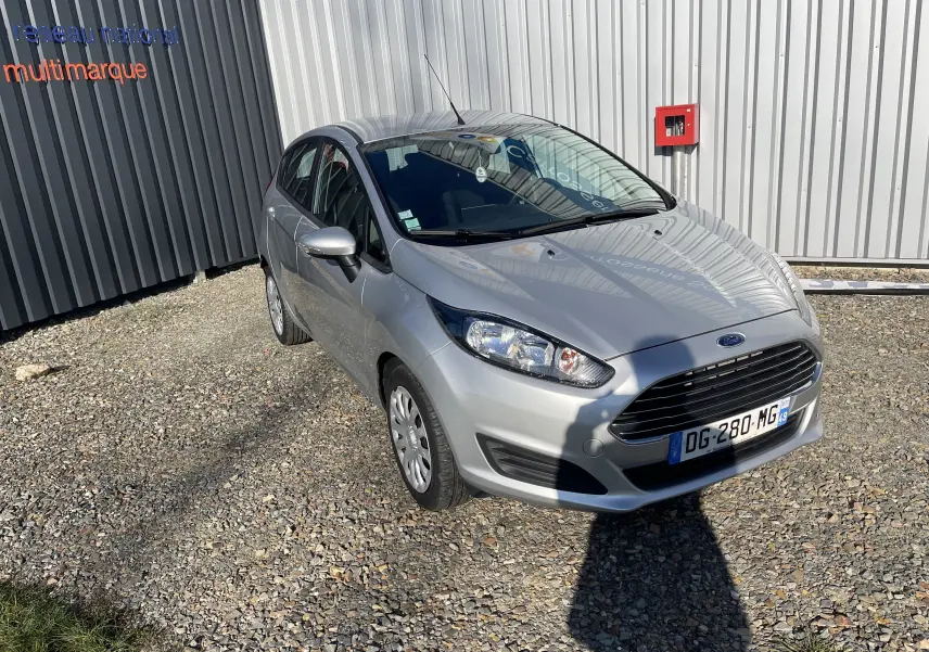 Ford Fiesta gris titane vue en 3/4 avant droit, garée sur sol gravillonné devant un mur métallique.