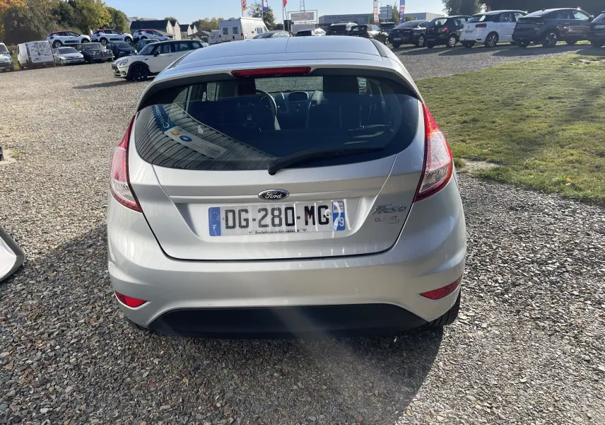 Vue arrière d'une Ford Fiesta gris titane 2014, avec plaque d'immatriculation française visible sur un terrain gravillonné.