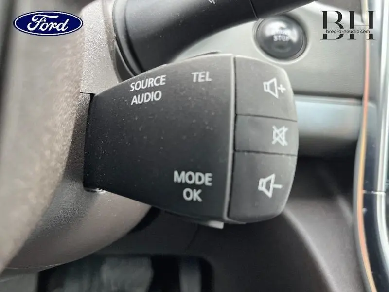 Gros plan sur la commande audio au volant d’un véhicule, avec boutons de volume et mode, intérieur noir.