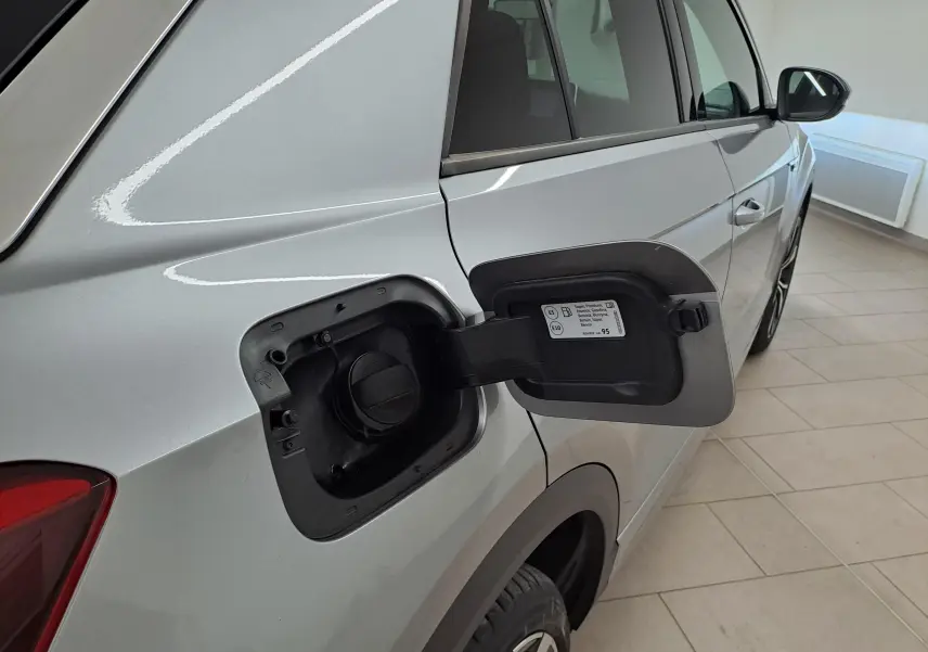 Vue arrière côté gauche du Volkswagen T-Roc gris pyrite avec trappe à carburant ouverte sur un sol carrelé intérieur.