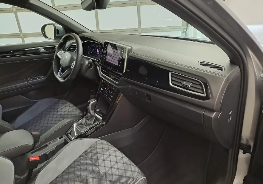 Vue intérieure côté conducteur du Volkswagen T-Roc 2025, avec tableau de bord noir brillant et sièges tissu à motif géométrique.