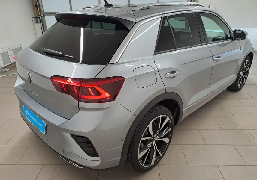 Vue 3/4 arrière droite du Volkswagen T-Roc gris pyrite avec jantes noires 19 pouces et feux arrière allumés.