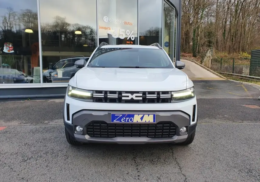 Vue avant d'un Dacia Duster blanc glacier 2025 avec calandre noire et feux LED allumés, stationné devant une vitrine.