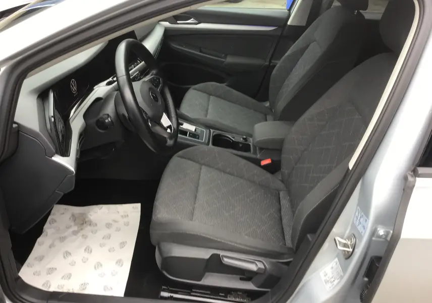 Vue intérieure côté conducteur de la Volkswagen Golf VIII gris reflex, montrant les sièges avant et le tableau de bord moderne.